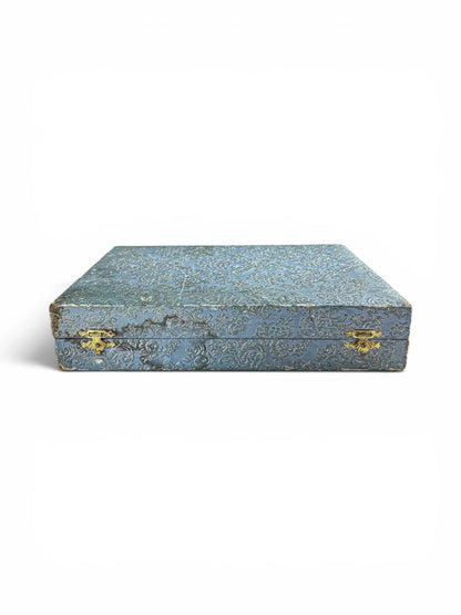 Front view of a vintage closed blue embossed damask presentation case for cutlery, showing age-authentic wear along the edges and two vintage brass-toned latches.