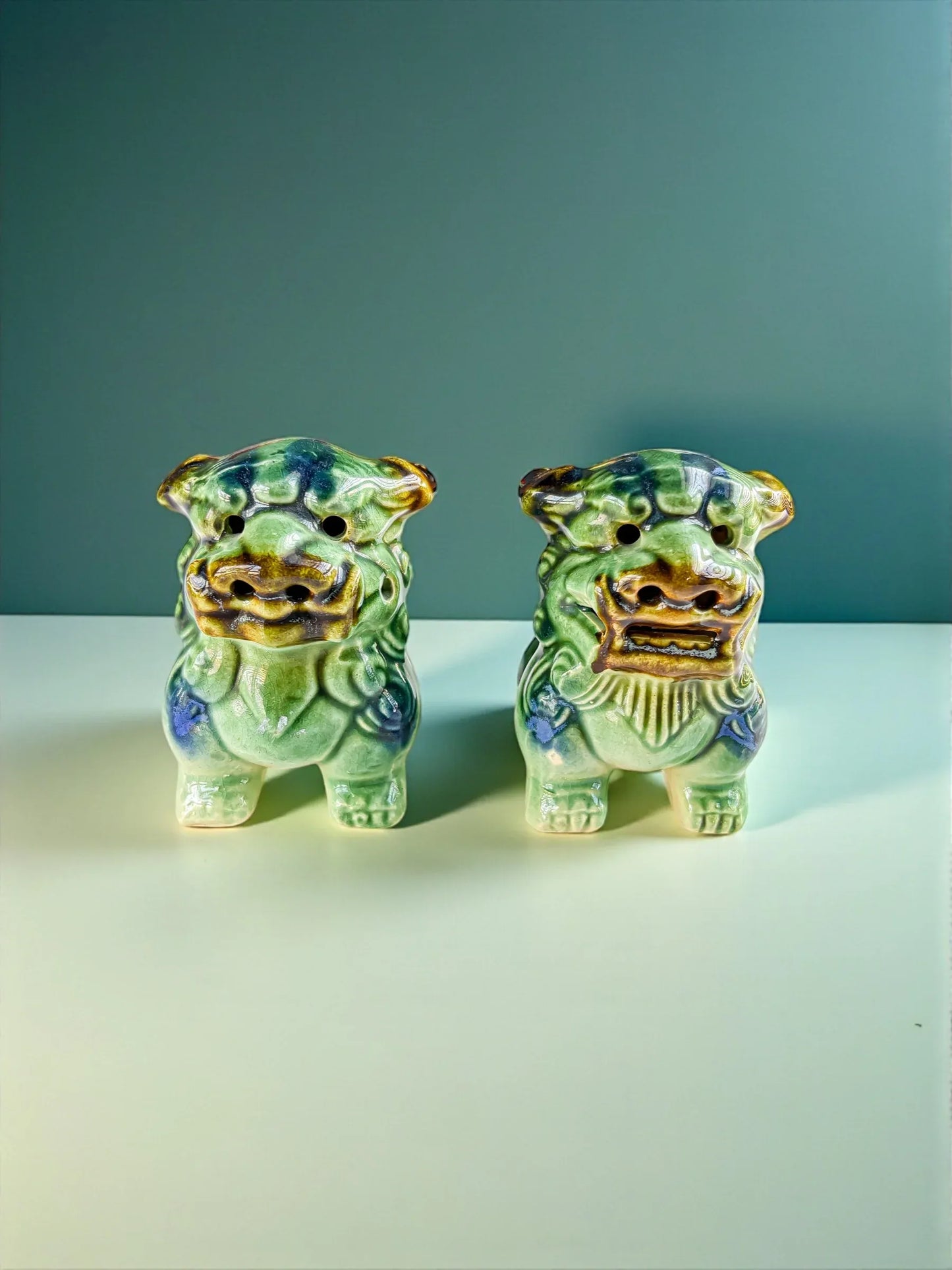 Pair of green ceramic Shisa lion statues with blue accents on white surface against teal background