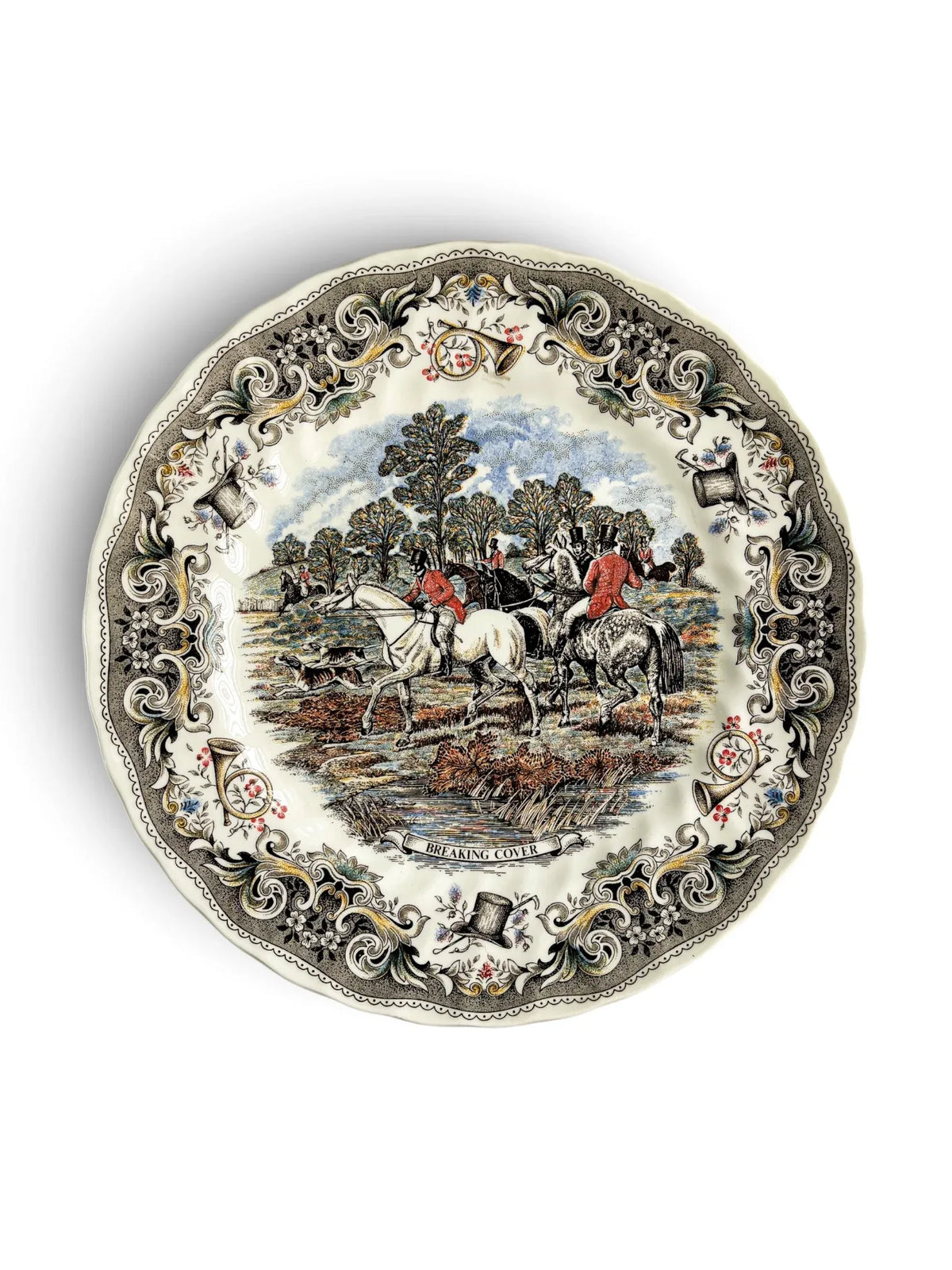Vintage Churchill Herring’s Hunt dinner plate titled Breaking Cover, showing riders in red coats on a white horse and a grey horse with hounds running left, water and reeds in the foreground, and an ornate grey border of horns, top hats and flowers. Scalloped rim. Photographed from above on a white background.