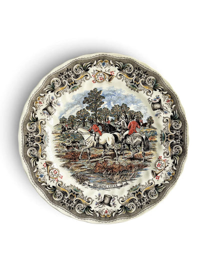 Vintage Churchill Herring’s Hunt dinner plate titled Breaking Cover, showing riders in red coats on a white horse and a grey horse with hounds running left, water and reeds in the foreground, and an ornate grey border of horns, top hats and flowers. Scalloped rim. Photographed from above on a white background.