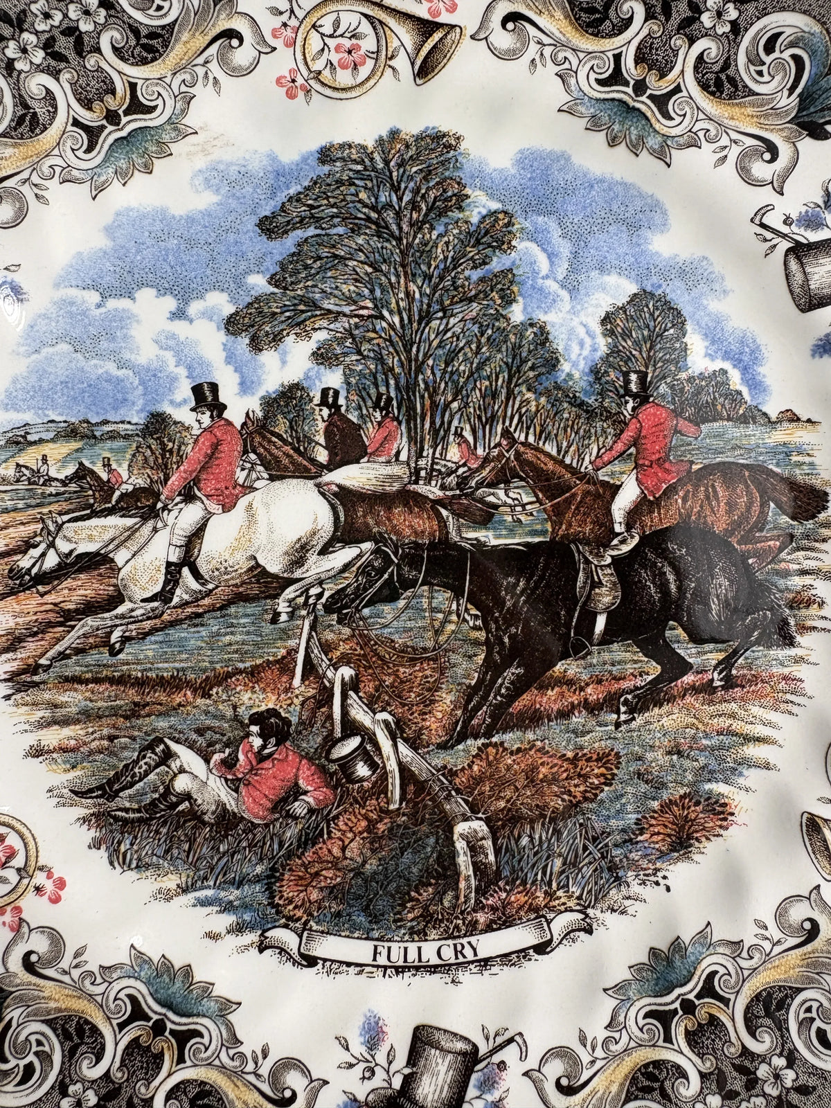 Close-up of the Full Cry transfer on a Churchill plate: hunters on a white horse, a brown horse and a black horse leaping a wooden fence, a rider lying on the ground in a red coat, trees and blue sky behind, with the title banner Full Cry at the bottom.