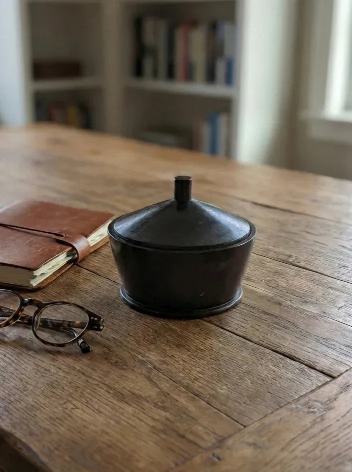 A minimalist black wooden lidded box sitting on a rustic wooden desk, styled next to a brown leather journal and tortoiseshell glasses.