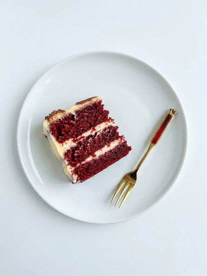 A flat lay photograph capturing a single gold-plated Eetrite cake fork with a ruby red handle, resting on a clean white saucer next to a simple white ceramic cup filled with dark tea, set against a smooth white background.