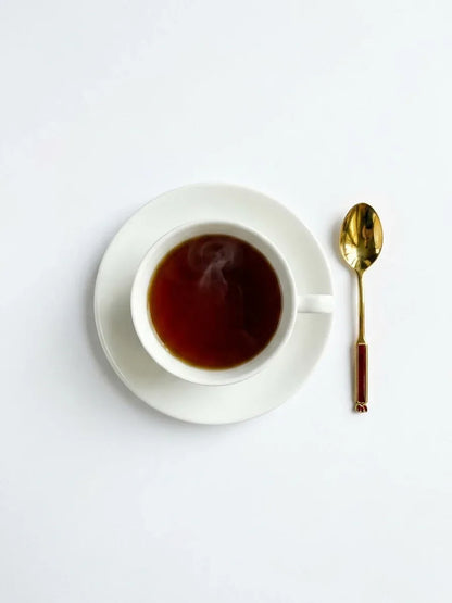 Flat lay overhead view of a single gold-plated Eetrite teaspoon with a red enamel handle resting next to a white ceramic teacup filled with dark tea on a white saucer.