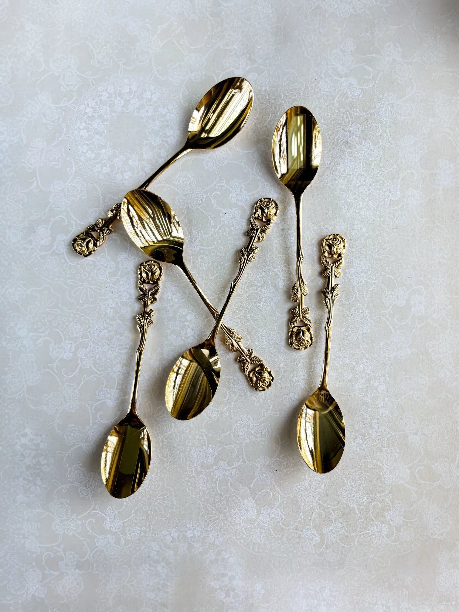 Set of six ornate gold spoons with rose-patterned handles on a light floral tablecloth
