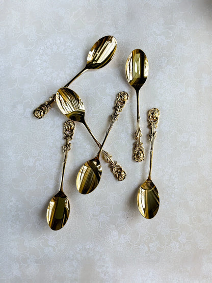 Set of six ornate gold spoons with rose-patterned handles on a light floral tablecloth