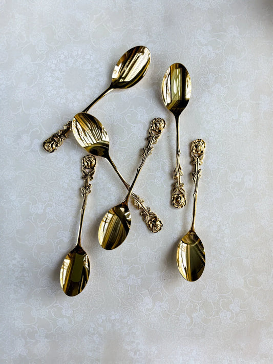 Set of six ornate gold spoons with rose-patterned handles on a light floral tablecloth