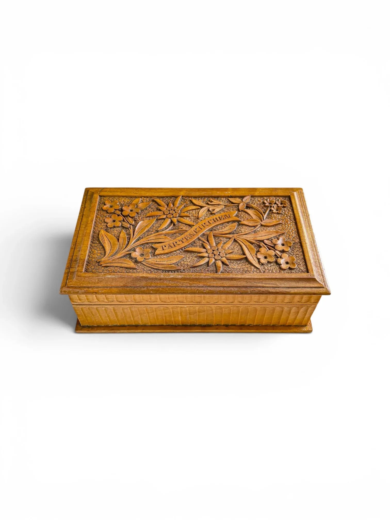 Top-down detail of a Bavarian wood-carved box lid showing intricate Edelweiss and Gentian motifs with a hand-carved ribbon bearing the inscription "Partenkirchen" over a stippled background.