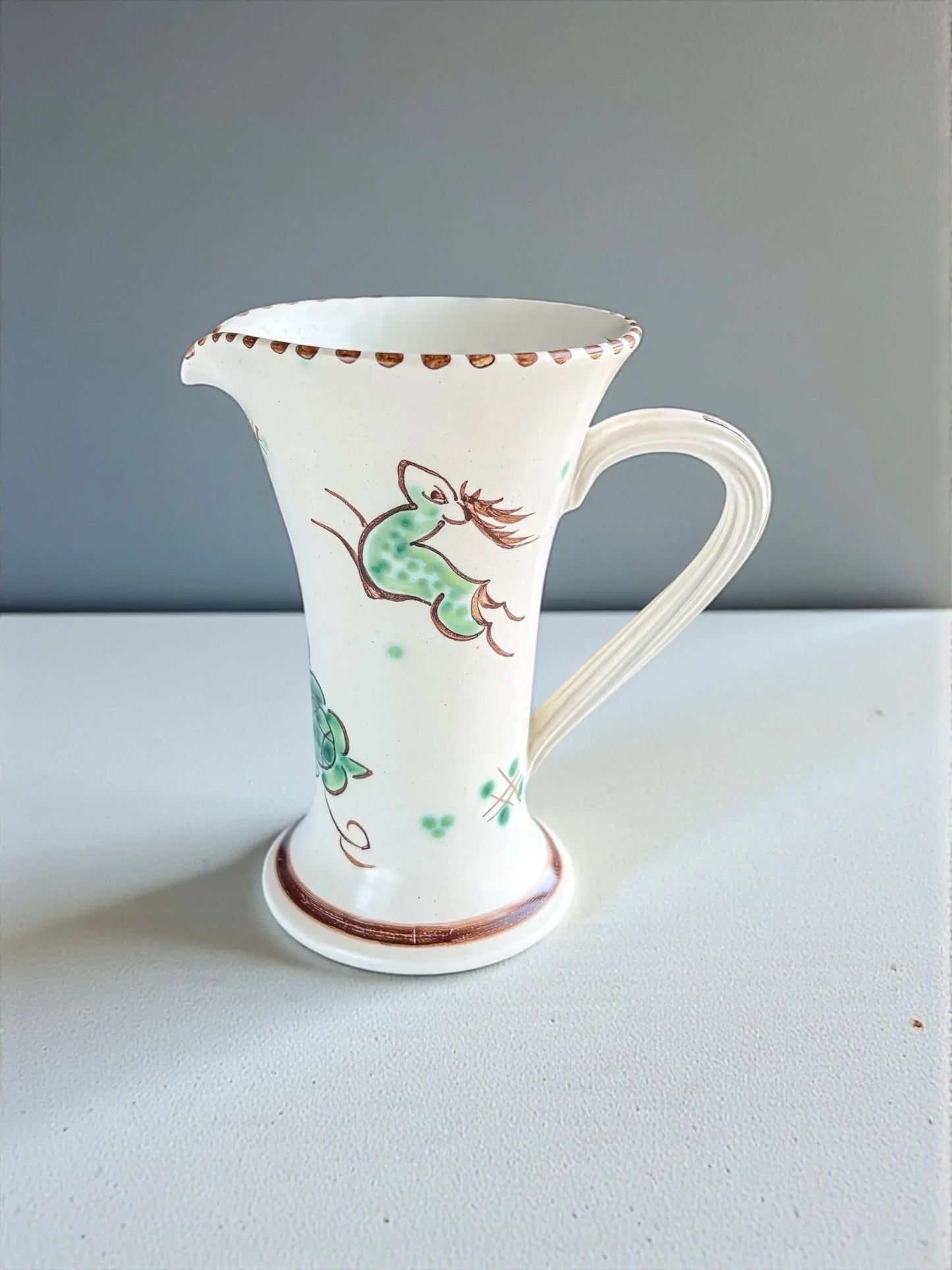 White ceramic pitcher with green and brown floral and bird design on white surface and light gray background