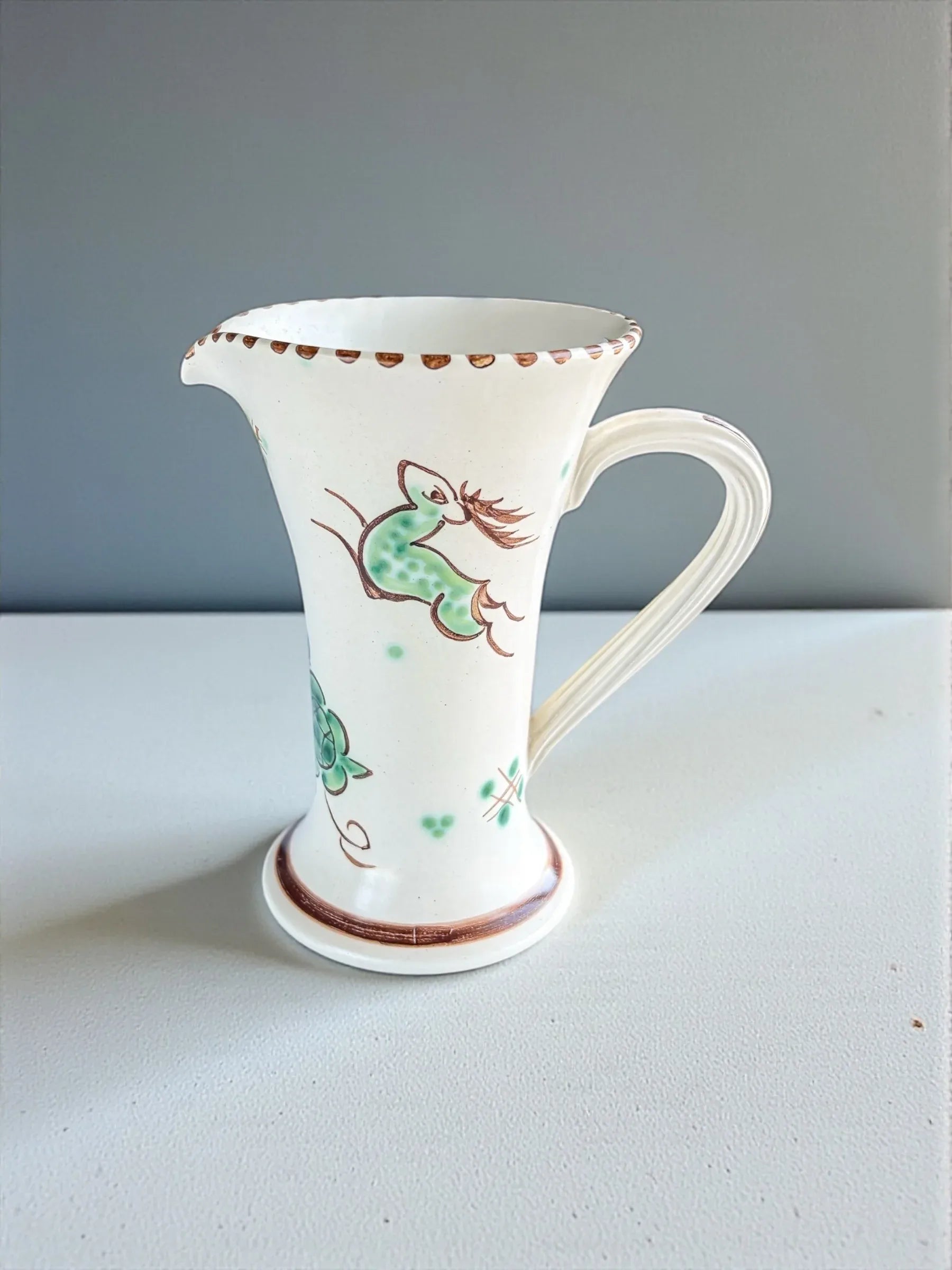 White ceramic pitcher with green and brown floral and bird design on white surface and light gray background