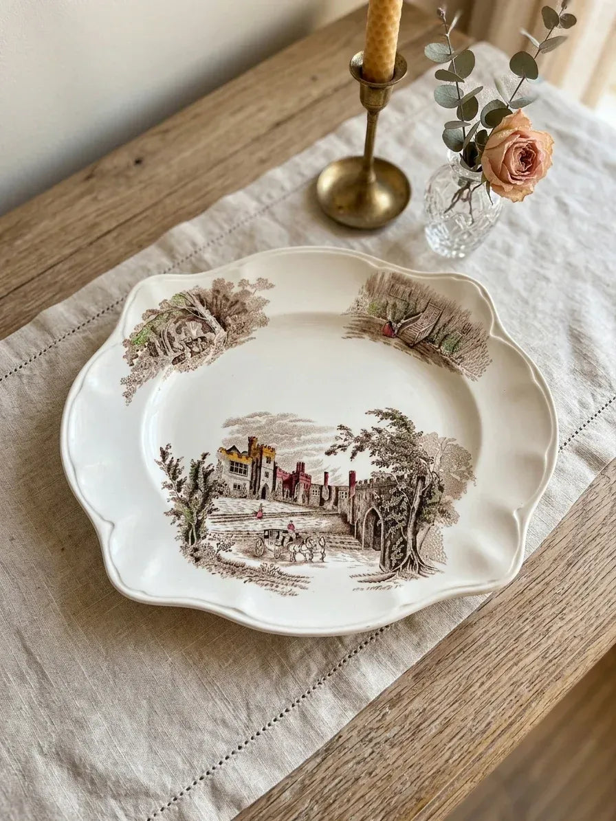 A vintage ceramic plate featuring a detailed brown transferware scene of a historic manor and horse-drawn carriage. The plate is styled on a neutral linen runner atop a rustic oak table, accompanied by a small crystal bud vase with a dried rose and a brass candlestick, creating a warm and sophisticated heritage aesthetic.