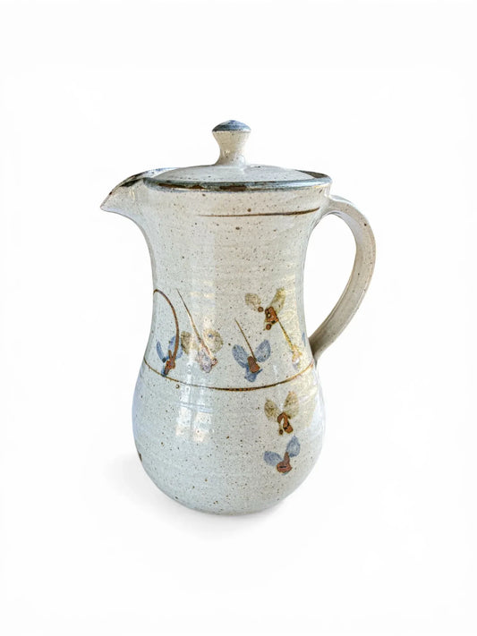 A front-facing view of a tall, lidded Kolonyama Pottery stoneware server featuring hand-painted cornflower blue and iron-oxide botanical motifs on a speckled oatmeal glaze. The piece has a tapered silhouette and a small, blue-tipped lid finial.