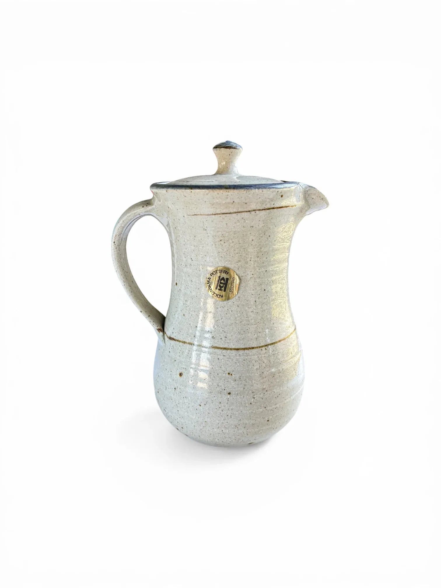 A side view of the lidded ceramic server showing the original circular gold foil merchant’s label, which reads "Kolonyama Pottery Lesotho." This angle displays the ergonomic hand-pulled handle and the subtle throwing rings of the studio pottery.
