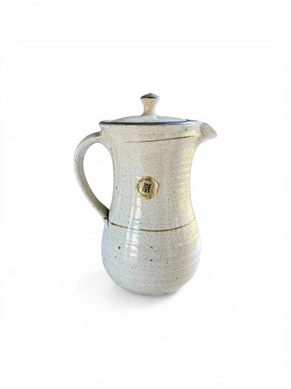 A side view of the lidded ceramic server showing the original circular gold foil merchant’s label, which reads "Kolonyama Pottery Lesotho." This angle displays the ergonomic hand-pulled handle and the subtle throwing rings of the studio pottery.