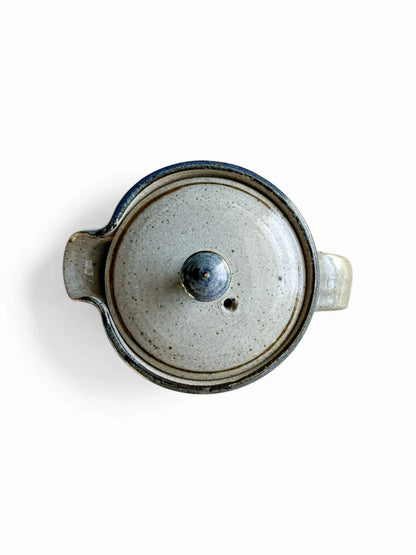 A detailed top-down photograph of the Kolonyama lidded server with the lid removed, revealing the clean, glazed internal cavity of the stoneware. The fitted lid sits beside the rim, showcasing the subtle blue rim detail and the pristine condition of the ceramic joins.