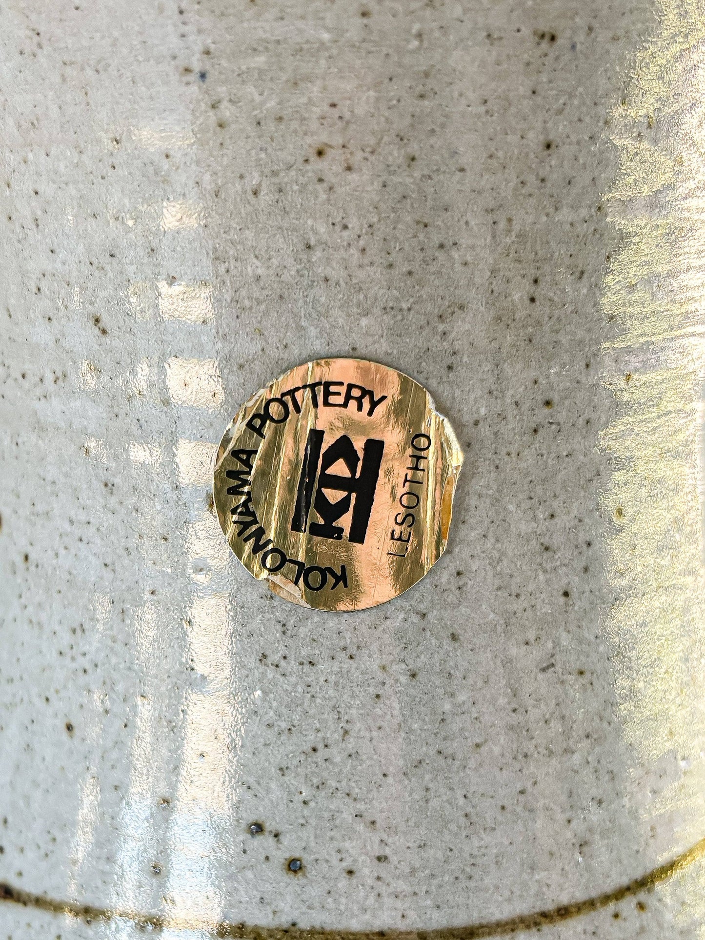 A high-resolution close-up focusing on the original circular gold foil sticker affixed to the side of the stoneware jug. The label remains predominantly intact and legible, featuring the 'K' logo and the text "KOLONYAMA POTTERY LESOTHO."
