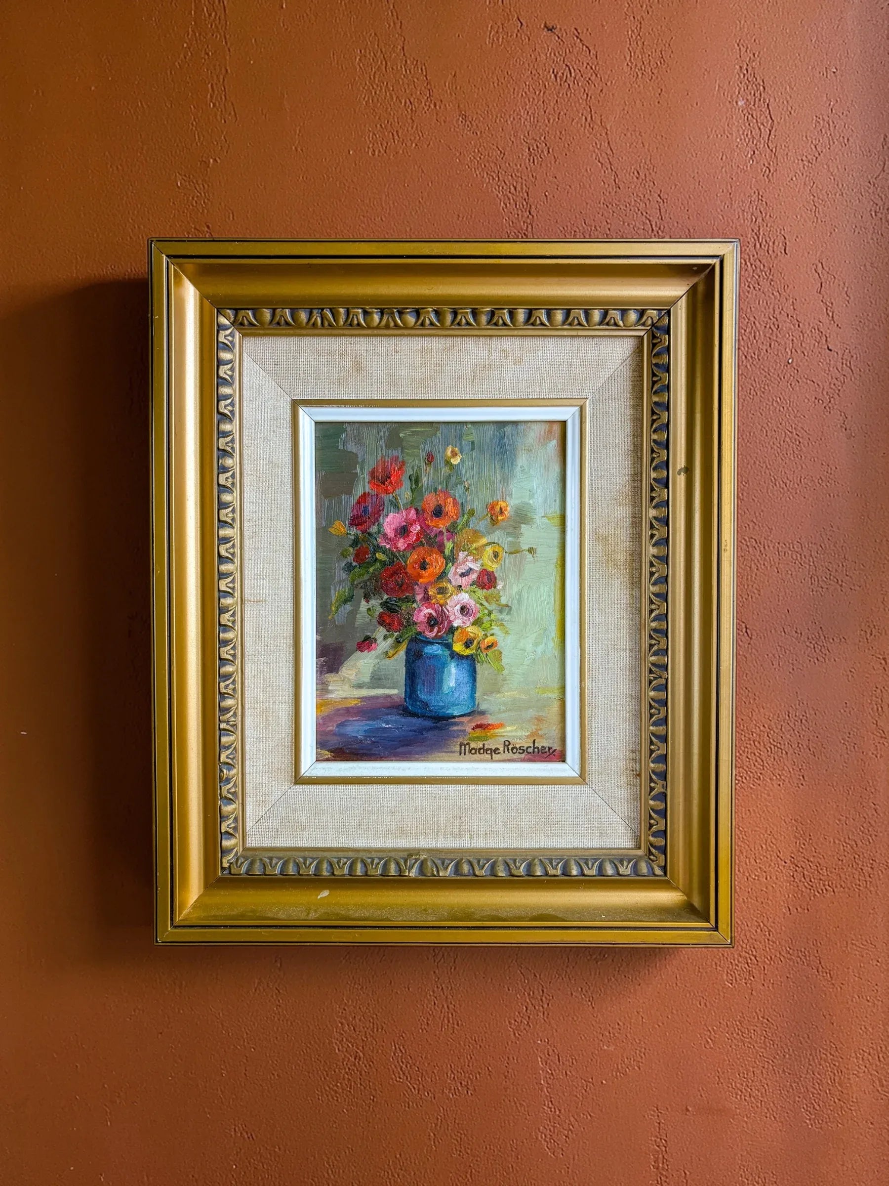A mid-century oil painting by Madge Röscher featuring a vibrant floral still life, displayed in a deep gold cove frame against a textured rust-orange wall.