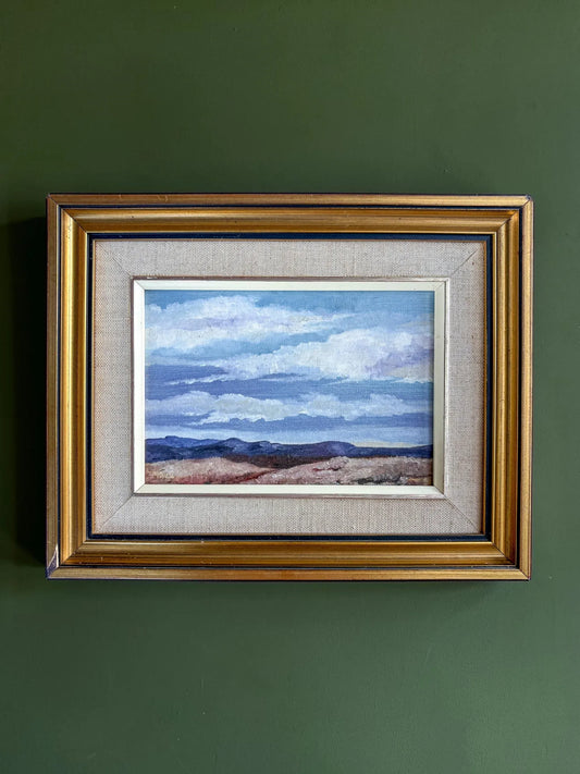 Framed vintage South African landscape oil painting hanging on a smooth, matte olive green wall. The gold gilt frame and textured beige linen slip stand out sharply against the deep green background.