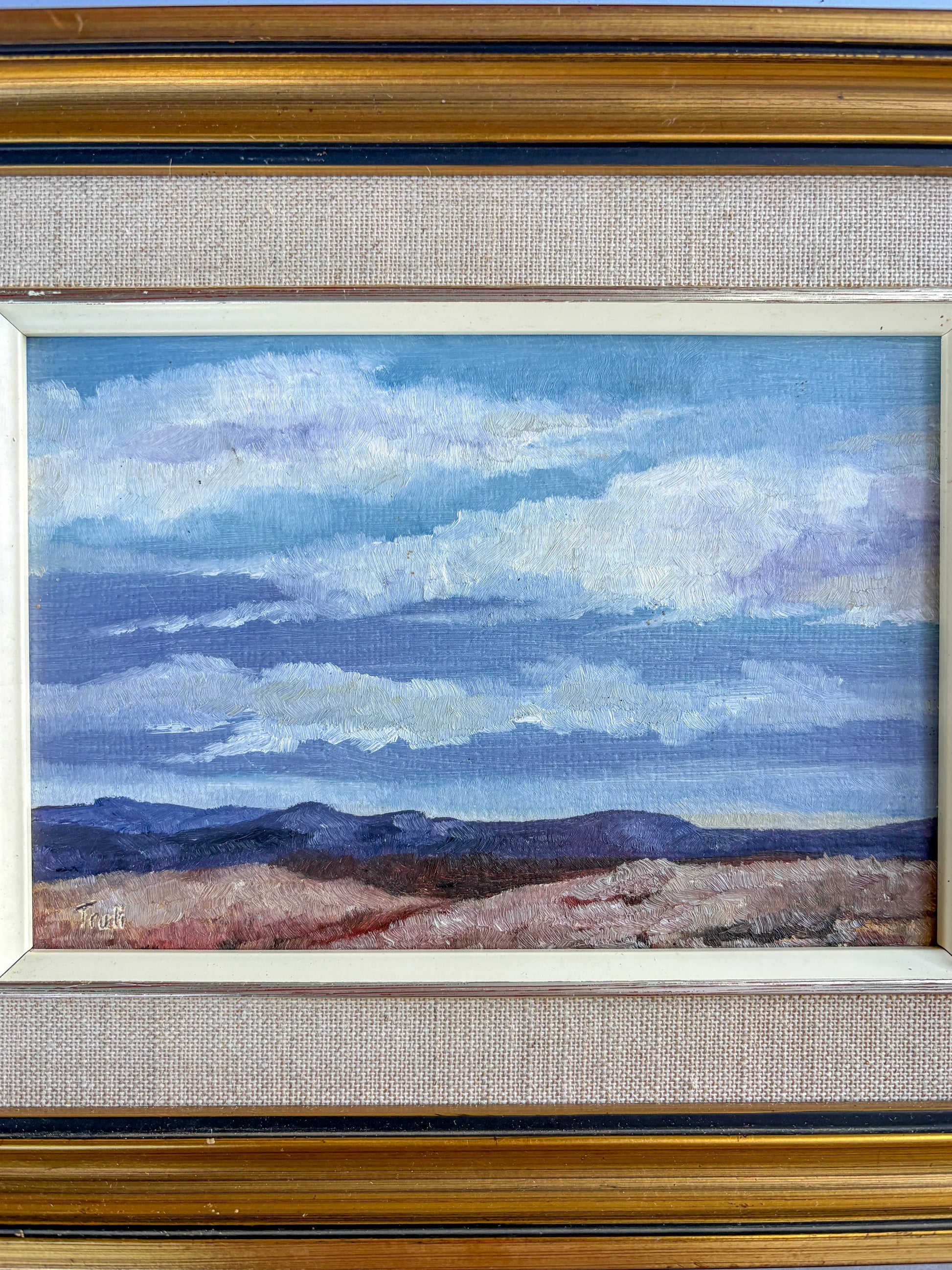 Close-up view of a vintage oil painting depicting a vast sky with layered white and blue clouds over distant purple hills and textured beige terrain. The detailed linen slip, white inner liner, and artist's signature in the corner are clearly defined.