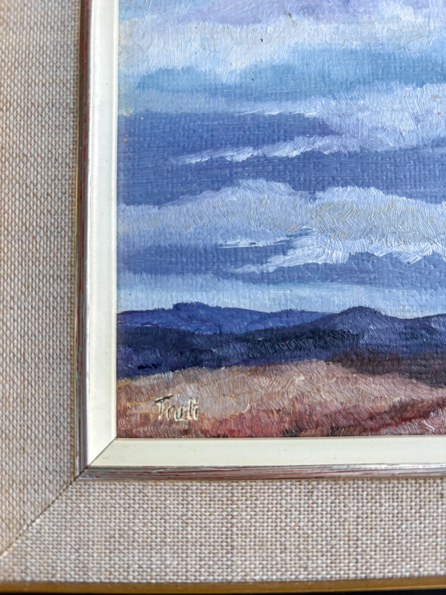 A detailed macro photograph highlighting the lower left corner of a vintage oil painting. The artist's handwritten signature is visible against the textured terrain foreground, framed by a slender cream inner border and a woven beige linen mat.