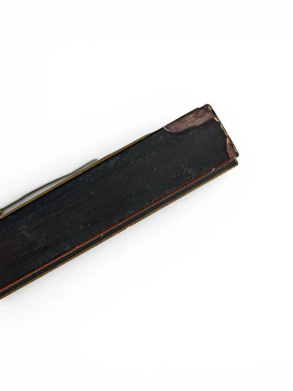 A close-up profile shot of the long side of a vintage wooden frame profile isolated on a clean white background. The dark, distressed lacquer backing is visible alongside thin gold and red trim lines along the decorative front lip.