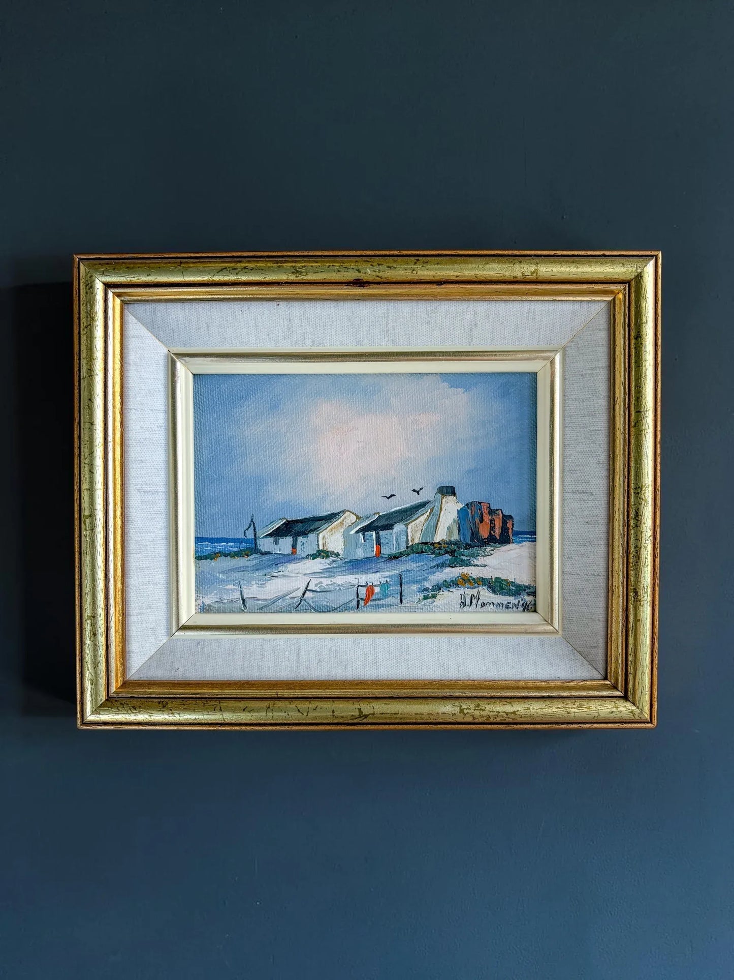 A framed original oil landscape painting by H. Mommen hanging on a dark matte navy blue wall. The artwork depicts traditional white-walled coastal fisherman cottages under a soft blue sky. It is enclosed in a layered frame featuring a light grey linen-textured slip and an outer gilt gold frame.