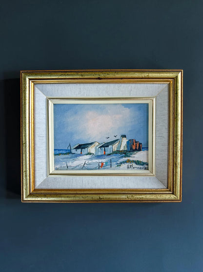 A framed original oil landscape painting by H. Mommen hanging on a dark matte navy blue wall. The artwork depicts traditional white-walled coastal fisherman cottages under a soft blue sky. It is enclosed in a layered frame featuring a light grey linen-textured slip and an outer gilt gold frame.