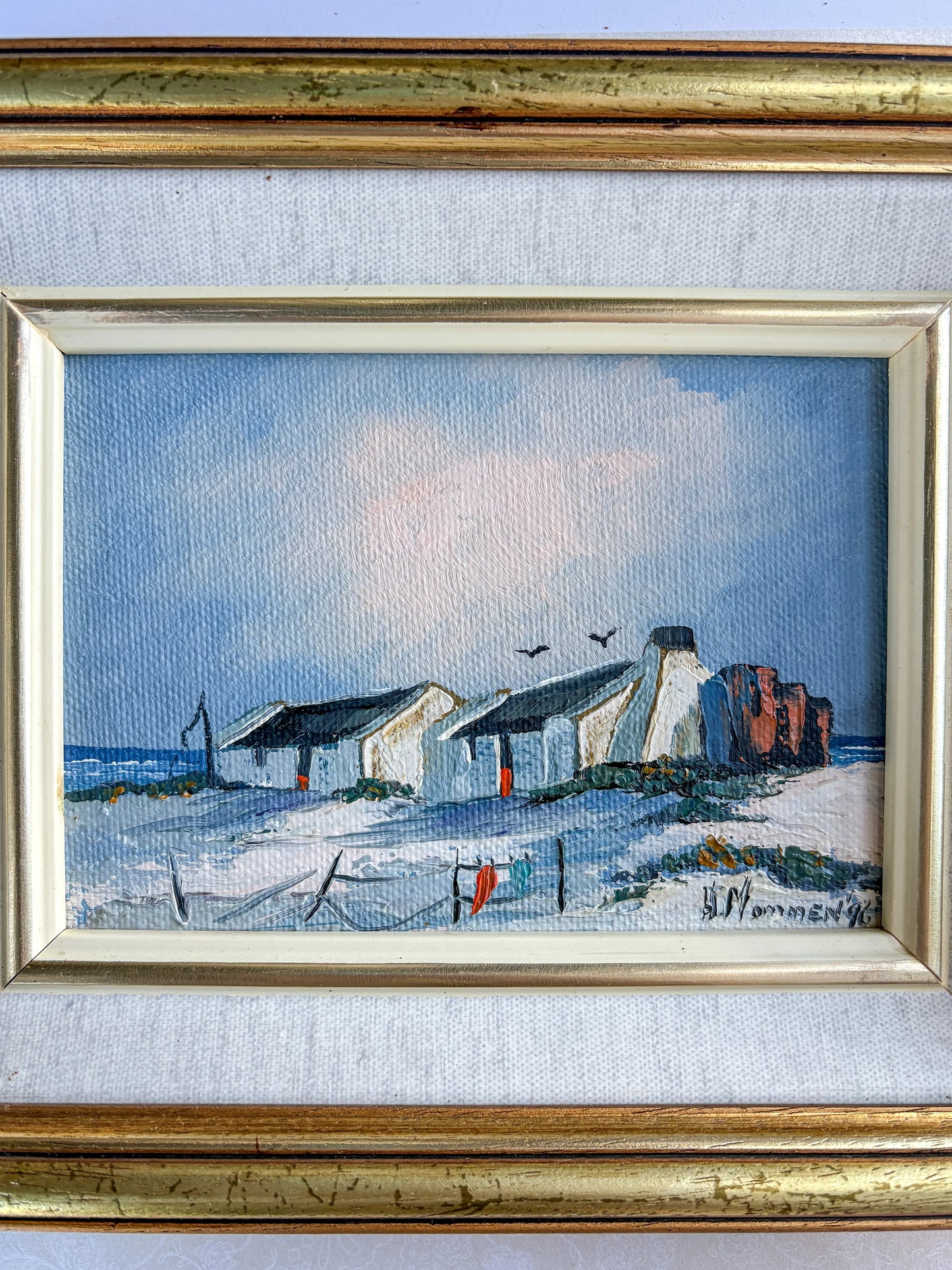 A close-up view focusing directly on the canvas window of H. Mommen's 1996 coastal painting. The detailed shot highlights the thick impasto texture of the whitewashed cottage walls, the sand dunes, and the vibrant red-orange pop of the cottage door and laundry hanging on a line.