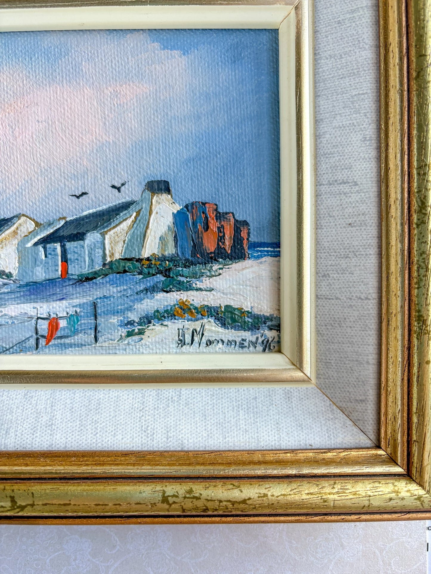 A cropped, detailed shot of the bottom right corner of the coastal landscape painting. The focus clearly shows the artist's dark blue painted signature, "H. Mommen '96," set against the textured white sand dunes, with a clear view of the adjacent inner gilt trim and linen slip frame.