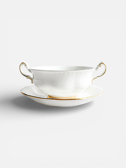 Elegant white porcelain soup bowl with gold-trimmed handles and matching saucer on white background