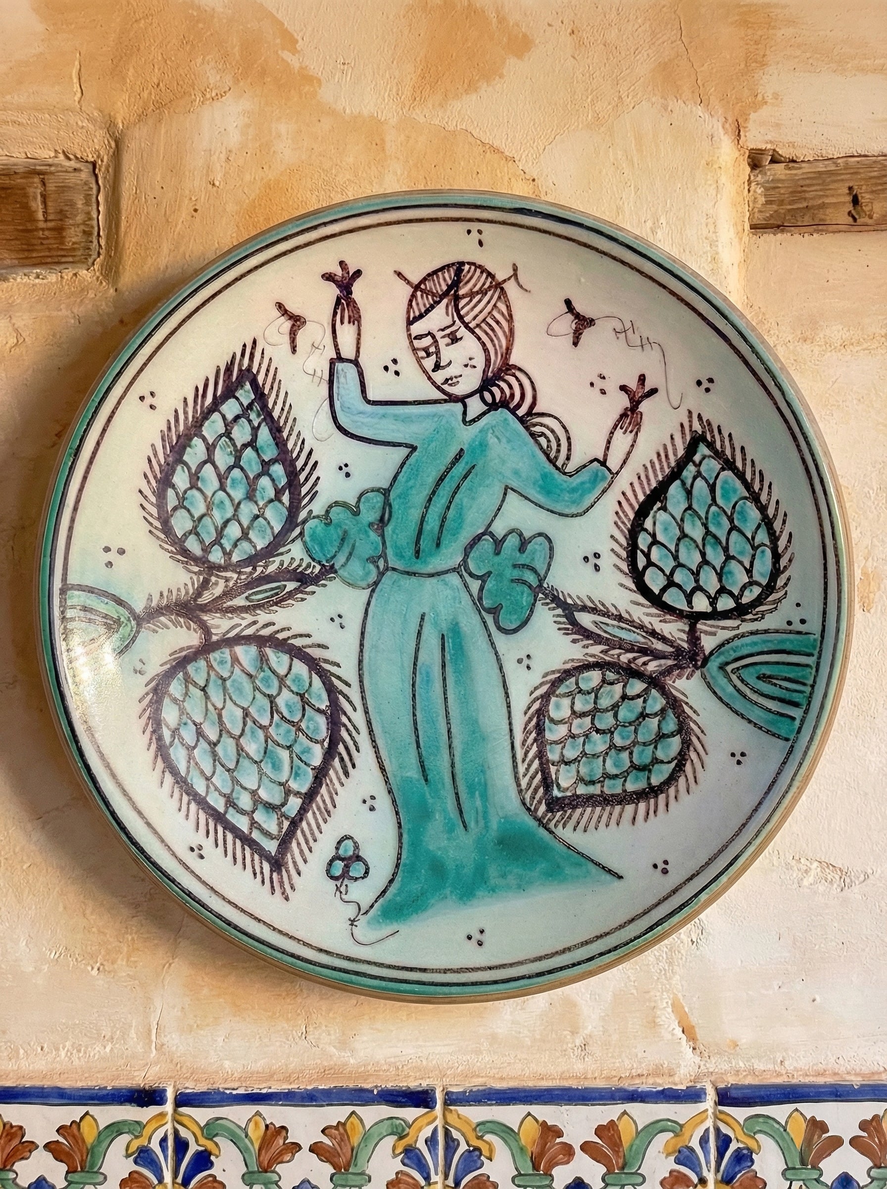 Decorative porcelain plate with a hand-painted figure in teal dress surrounded by floral motifs, displayed on a textured beige wall above colorful ceramic tiles