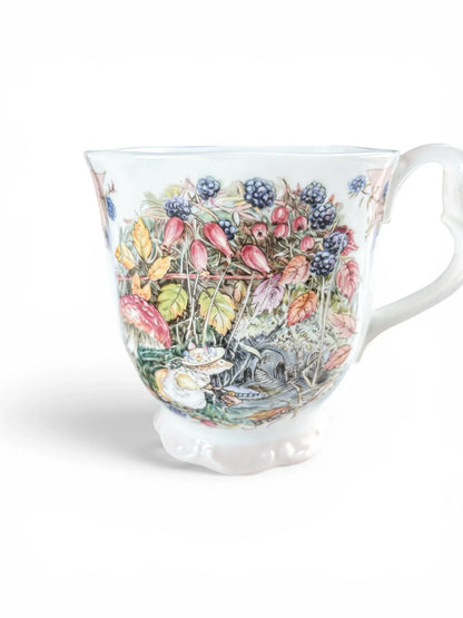 Close-up of the harvest illustration on a Royal Doulton Brambly Hedge Autumn beaker, highlighting the vibrant blackberries, autumn leaves, and the signature baby pink decorative ribbon along the rim.