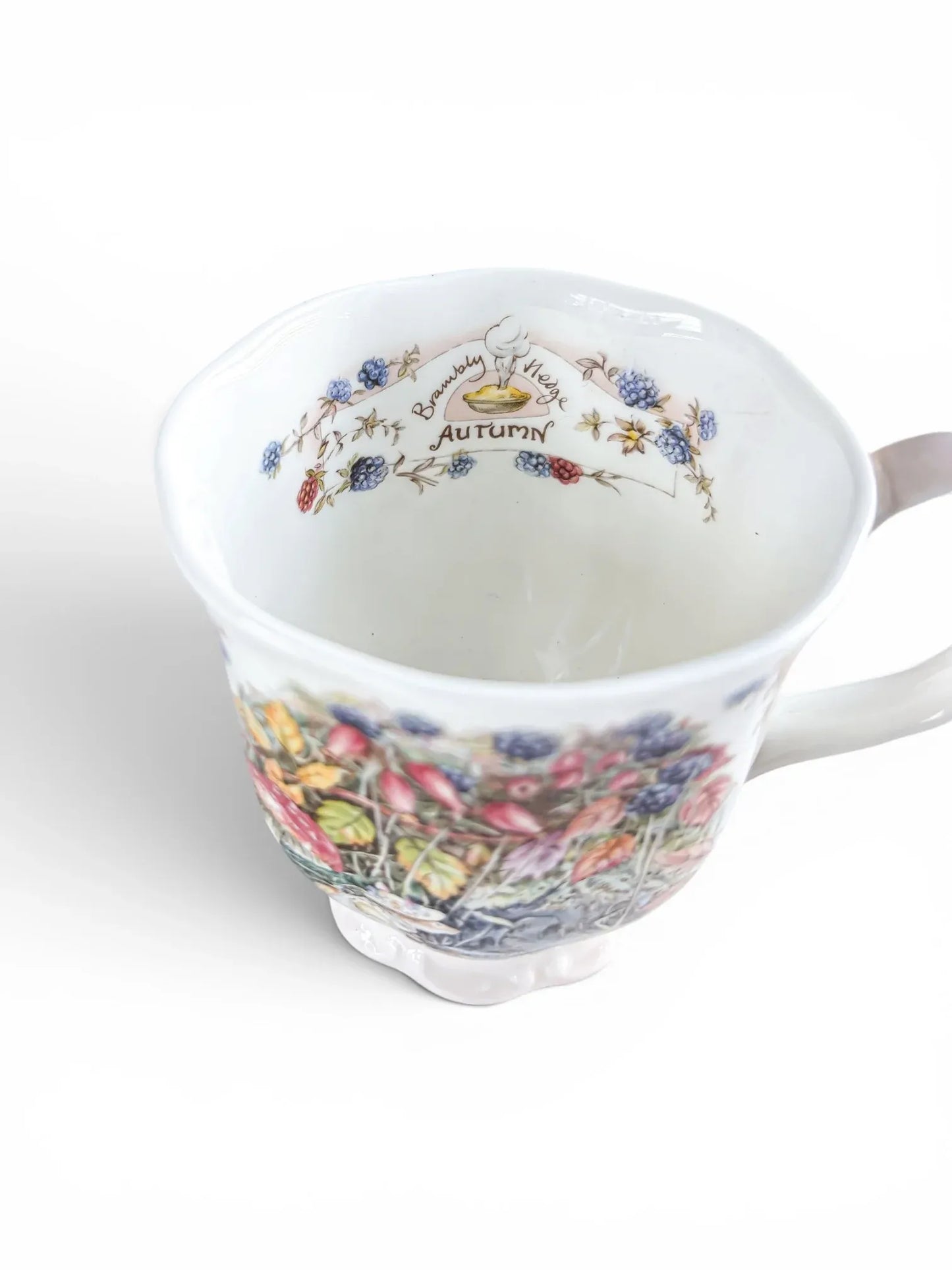 View looking into the interior of the Royal Doulton Brambly Hedge beaker, showcasing a hidden vignette on the inner rim of a steaming blackberry pie framed by an "Autumn" banner and bramble berries.