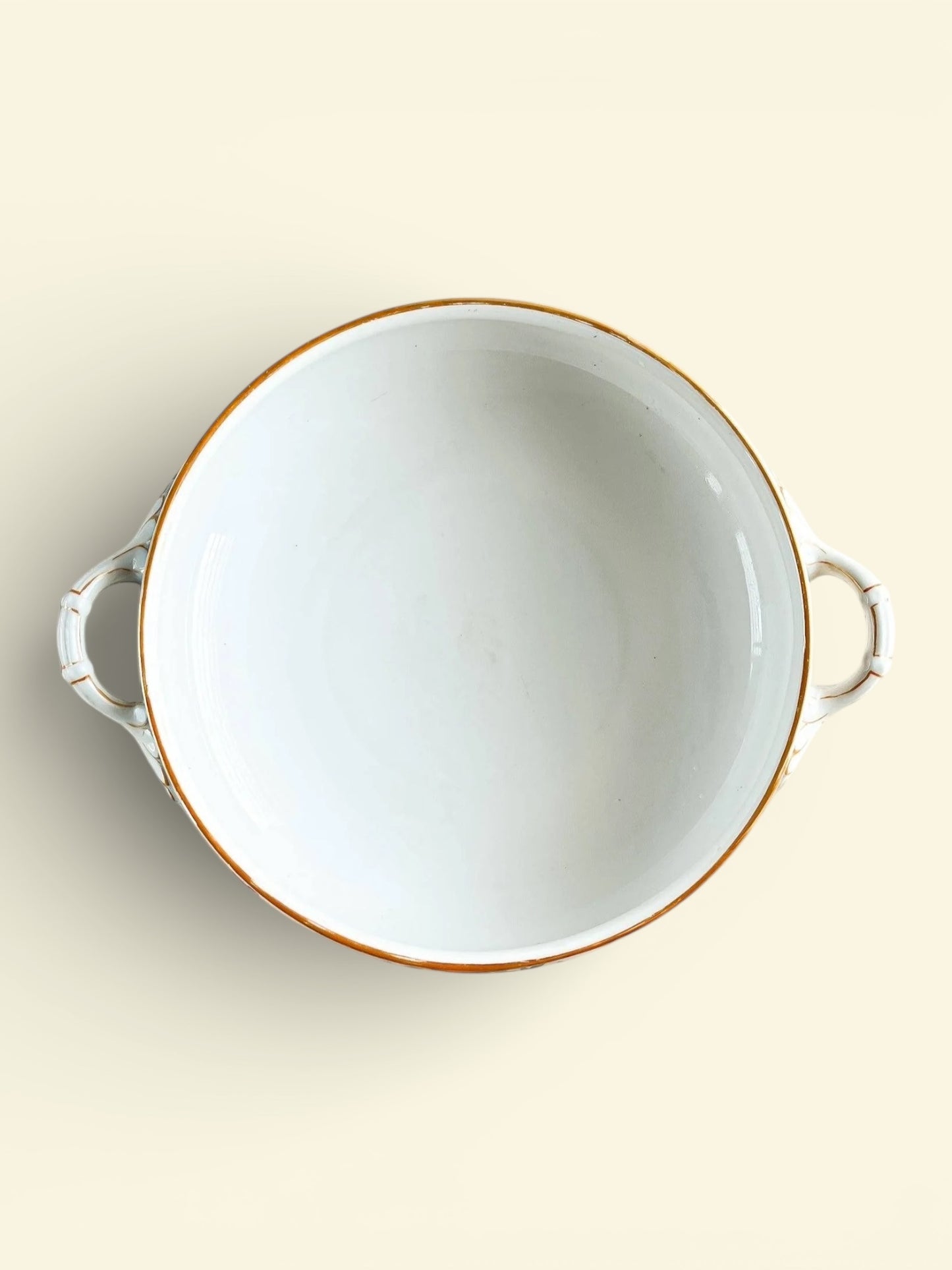 Top view of a white porcelain serving bowl with gold rim and two side handles on beige background