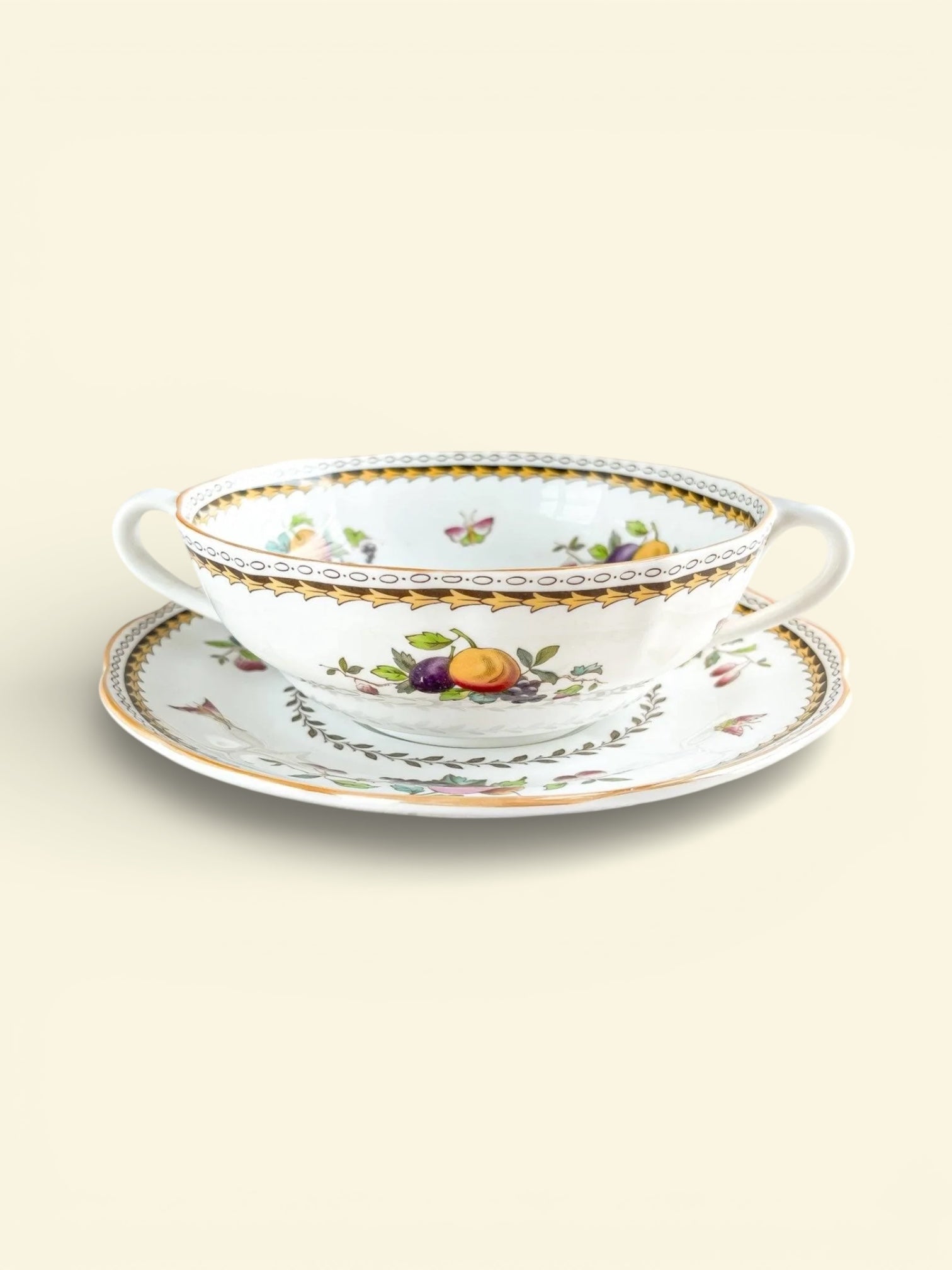 Vintage porcelain double-handled cup and saucer with gold trim and fruit floral pattern on soft beige background