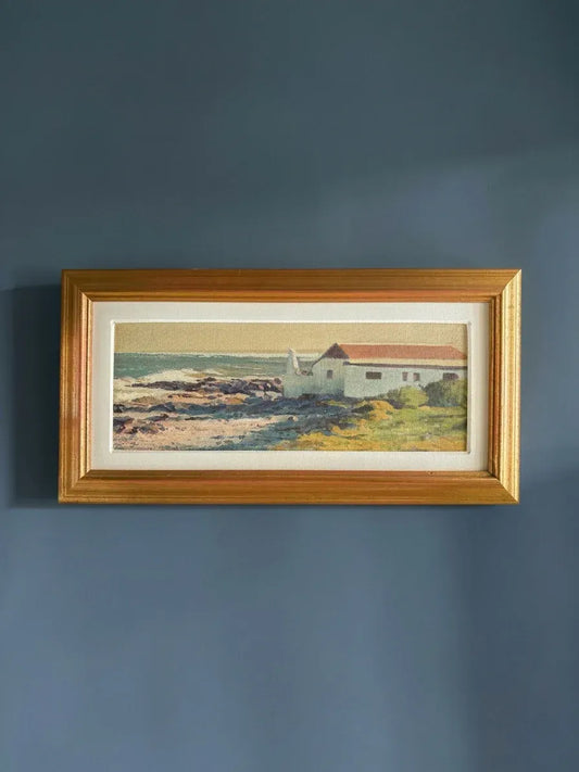 A panoramic Hannes Meintjies oil painting of a coastal fisherman's cottage in a vintage gold frame, styled against a moody deep blue wall with soft shadows.