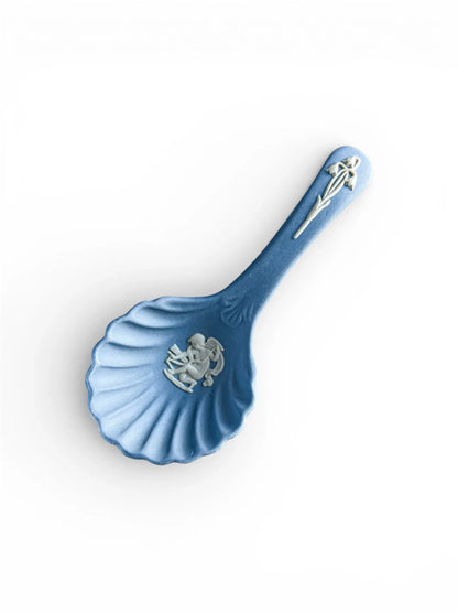 A top-down professional shot of a pale blue Wedgwood Jasperware caddy spoon on a clean white background. The scalloped shell-shaped bowl features a white bas-relief of Cupid sharpening his arrow, and the handle is adorned with a delicate white lily of the valley sprig.