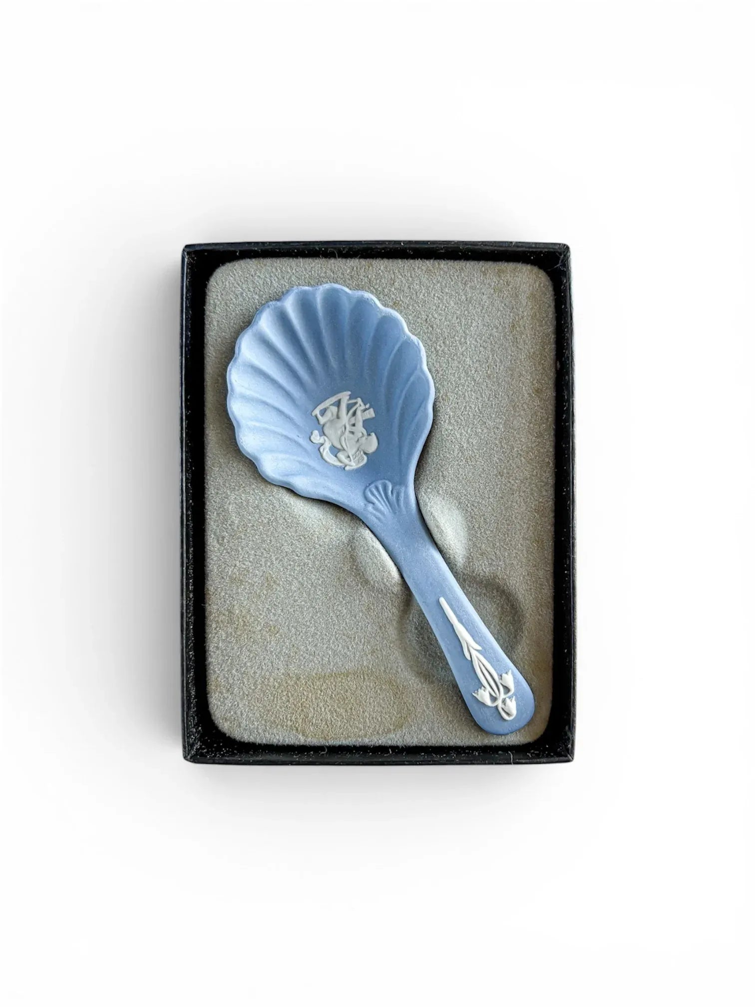 A pale blue Wedgwood Jasperware shell caddy spoon nestled in its original dark grey presentation box with a contoured foam insert. The white mythological Cupid scene and handle detail are clearly visible against the matte blue stoneware.