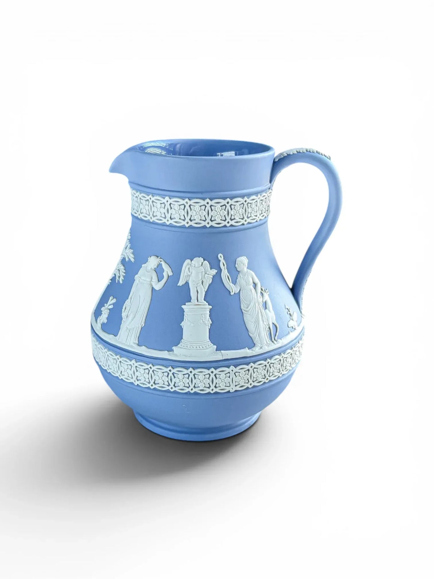 A vintage 1956 blue Wedgwood Jasperware Ejug with a glazed interior. The light blue matte stoneware body is decorated with white bas-relief classical figures, a small cupid on a pedestal, and detailed floral geometric borders. The classic loop handle features a white acanthus leaf motif, all shot against a clean white background.