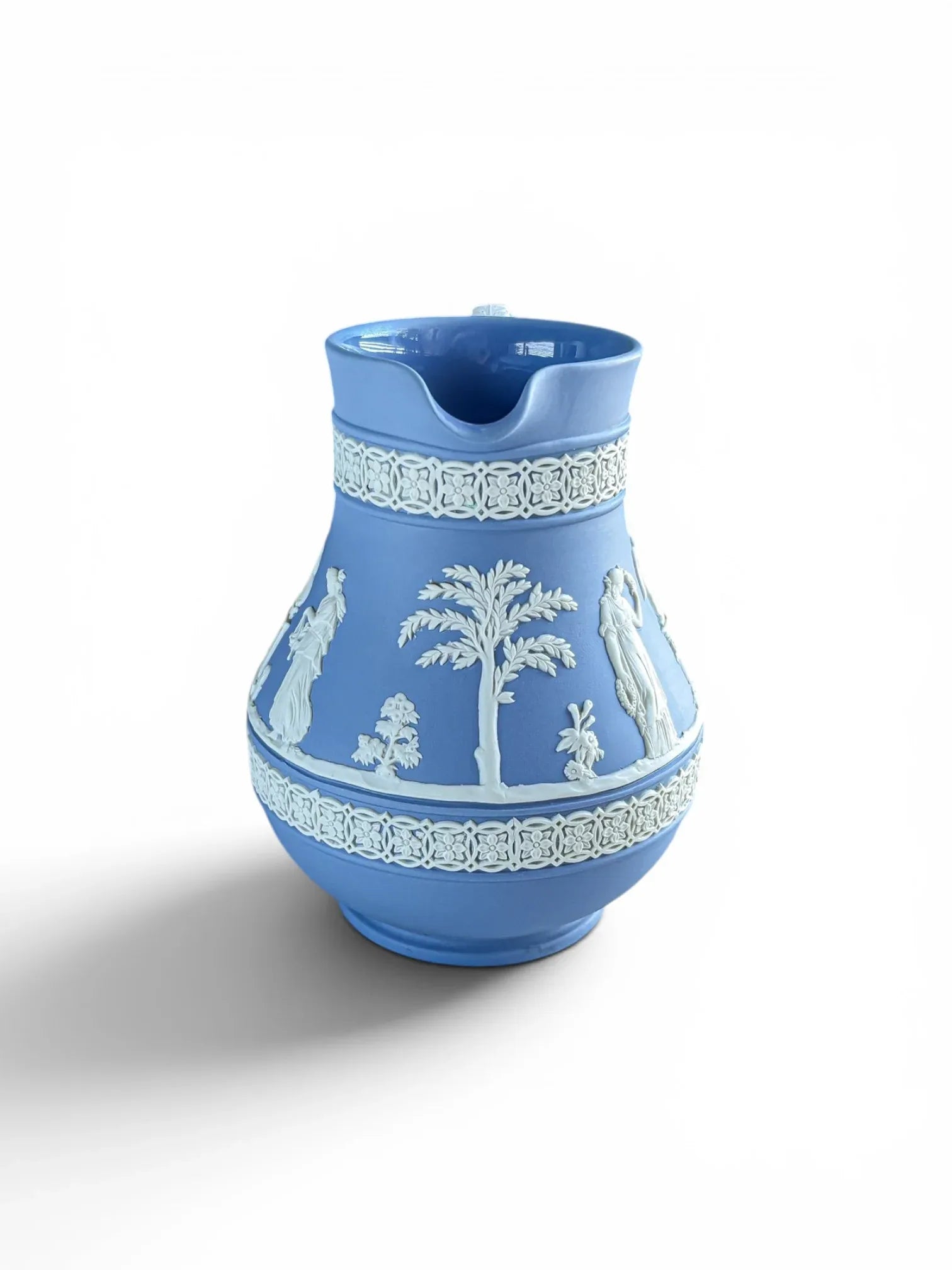 Front angle view of a vintage mid-century blue Wedgwood Jasperware Etruscan pitcher showing the pouring spout. The matte light blue porcelain body features intricate white sprigged reliefs of a classical palm tree and Roman figures, framed by matching white ornamental bands at the neck and base.