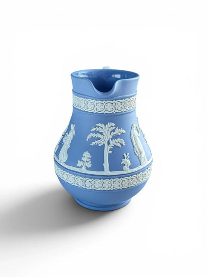 Front angle view of a vintage mid-century blue Wedgwood Jasperware Etruscan pitcher showing the pouring spout. The matte light blue porcelain body features intricate white sprigged reliefs of a classical palm tree and Roman figures, framed by matching white ornamental bands at the neck and base.