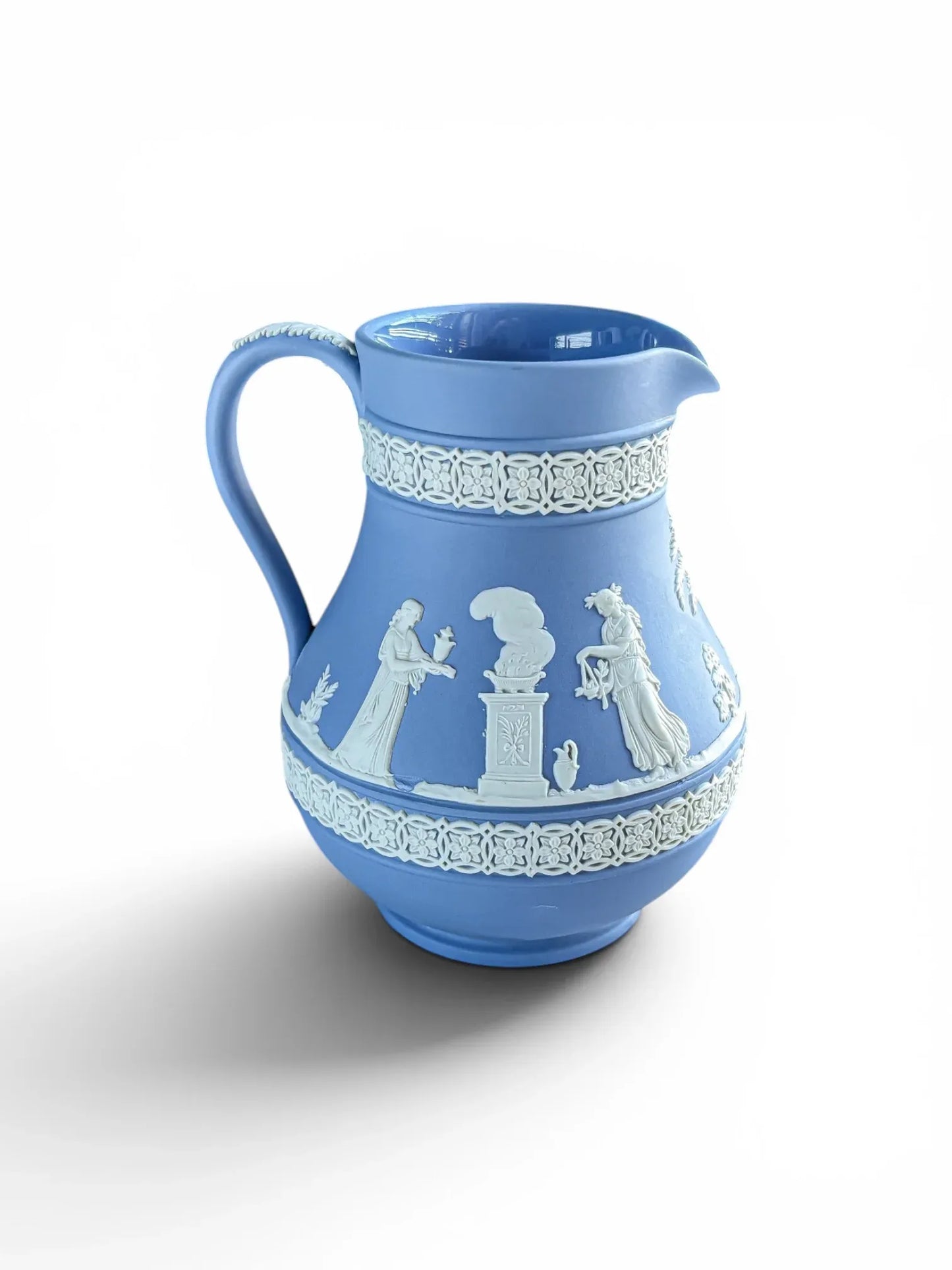 Side view of a 1956 vintage Wedgwood blue Jasperware Etruscan jug on a solid white background. This angle features a prominent white bas-relief scene depicting two classical Roman women offering sacrifices at a smoking altar pillar, framed by upper and lower geometric floral bands.