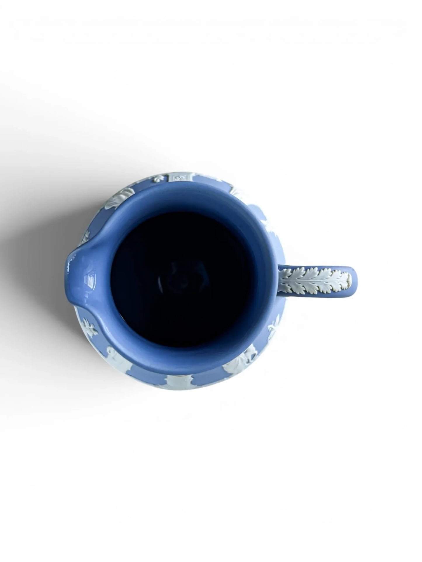 A top-down bird's-eye view of a vintage blue Wedgwood Jasperware Etruscan jug, clearly displaying the glossy, smooth glazed interior lining. The image shows the circular opening, the pouring lip to the left, and the white acanthus leaf relief detail along the top curve of the handle to the right.