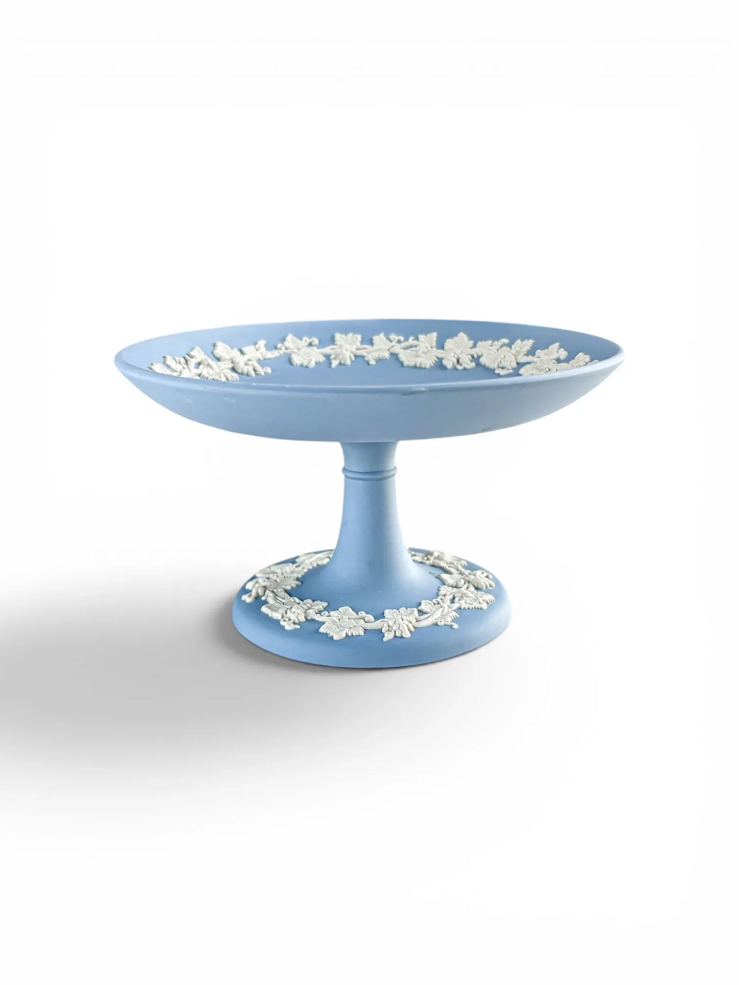 A professional profile shot of a pale blue Wedgwood Jasperware footed compote against a clean white background. The image highlights the elegant pedestal silhouette and the intricate white grapevine bas-relief border encircling both the interior rim and the circular base.