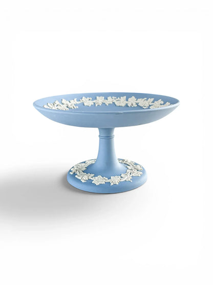 A professional profile shot of a pale blue Wedgwood Jasperware footed compote against a clean white background. The image highlights the elegant pedestal silhouette and the intricate white grapevine bas-relief border encircling both the interior rim and the circular base.