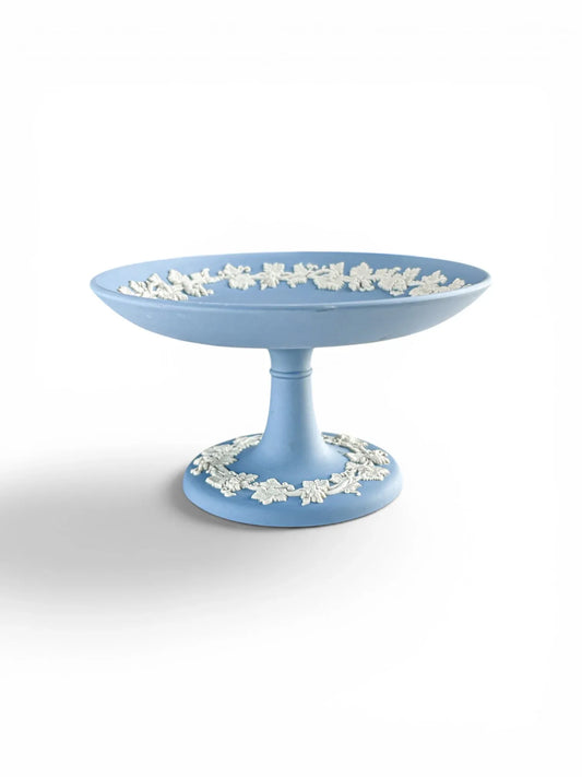 A professional profile shot of a pale blue Wedgwood Jasperware footed compote against a clean white background. The image highlights the elegant pedestal silhouette and the intricate white grapevine bas-relief border encircling both the interior rim and the circular base.