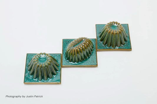An overhead perspective of three sculptural ceramic tiles on a white background. The tiles, featuring emerald green speckled bases and ribbed, amber-toned central domes, are positioned in a stepped diagonal arrangement to showcase the reflective glaze and geometric form.