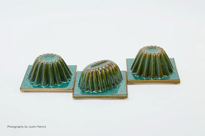 A professional studio shot against a stark white background featuring three sculptural ceramic tiles arranged in a diagonal line. Each square tile is topped with a fluted, jelly-mould-inspired dome finished in a high-gloss, variegated green glaze.