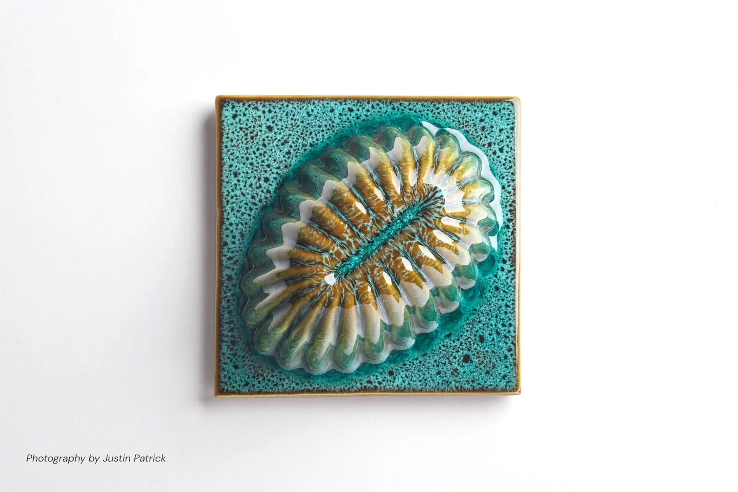A top-down view of a square-shaped ceramic sculptural tile. The central fluted motif is elongated into an oval shape, showcasing a rich crystalline glaze in shades of green and gold over a textured, teal-coloured square base.