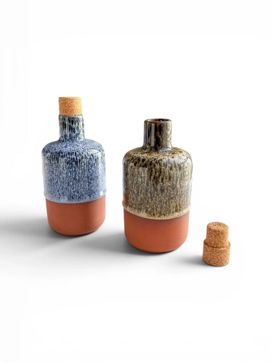 Two Wiid Design half-body ceramic olive oil bottles on a white background. One is finished in cobalt blue reactive glaze and the other in brown, both featuring raw terracotta bases and natural cork stoppers.